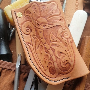 May include: A light brown leather sheath with a floral design, brass rivet, and black stitching. The sheath is surrounded by leatherworking tools, including a multi-tool, awls, and a wooden burnisher. The tools are for leathercraft.
