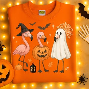 May include: An orange sweatshirt with a Halloween-themed design. It features three flamingo characters dressed as a witch, a jack-o'-lantern, and a ghost. The design also includes bats, stars, and a spiderweb, perfect for the spooky season.