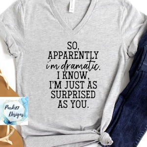 May include: A light gray V-neck t-shirt with the text "SO, APPARENTLY I'M DRAMATIC. I KNOW, I'M JUST AS SURPRISED AS YOU." in black. The shirt is paired with blue jeans and brown sandals.