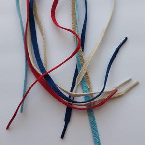 May include: A collection of colorful shoelaces in various shades of blue, red, and beige. The laces are flat and woven, with plastic aglets at the ends. The laces are arranged on a white background.