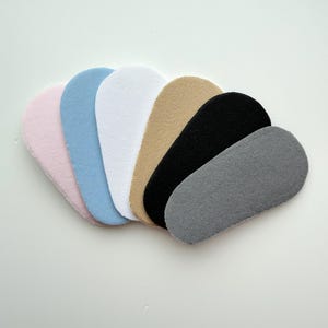 May include: A collection of seven oval-shaped shoe inserts in various colors, including pink, light blue, white, tan, black, and gray. The inserts have a textured surface, likely for added comfort and grip within footwear.