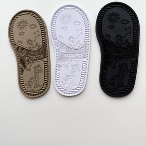 May include: Three rubber shoe soles in tan, white, and black. Each sole features a textured border, the number 21, the text TUTTI FRUTI, and embossed illustrations of a sun, clouds, a bird on a branch, and a giraffe standing under a rainbow.