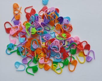 May include: A collection of colorful plastic stitch markers in various shades, including blue, green, orange, pink, purple, and red. These small, curved markers are used for knitting and crochet projects. The markers are scattered on a white surface.