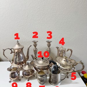 Könnte beinhalten: Eine Sammlung antiker silberner Tee- und Kaffeeservice-Artikel. Enthält Teekannen, einen Sahnekännchen, Zuckerdosen, eine Kaffeekanne und zwei Kerzenhalter. Die Artikel sind auf einer weißen Oberfläche angeordnet.