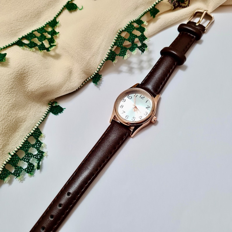 May include: A brown leather wristwatch with a white face and black numbers. The watch has a rose gold case and hands.