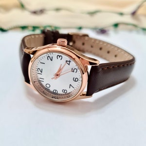 May include: A rose gold wristwatch with a white face and black numbers. The watch has a brown leather band.