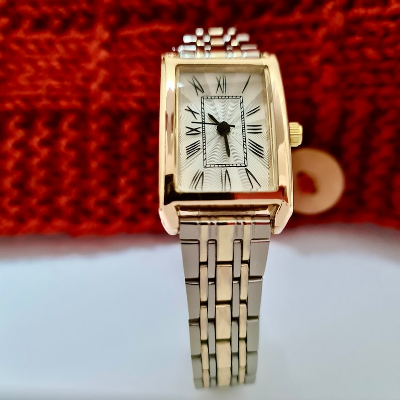 May include: A gold and silver toned watch with a rectangular face and Roman numeral hour markers. The watch has a white face with black hands and a silver band.