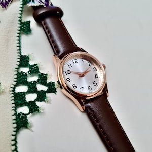 May include: A rose gold wristwatch with a white face and black numbers. The watch has a brown leather band.