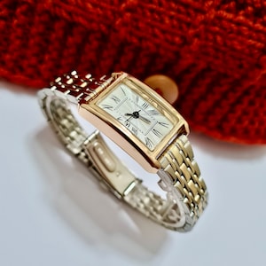 May include: A gold and silver toned watch with a rectangular face and Roman numeral hour markers. The watch has a two-tone metal band.