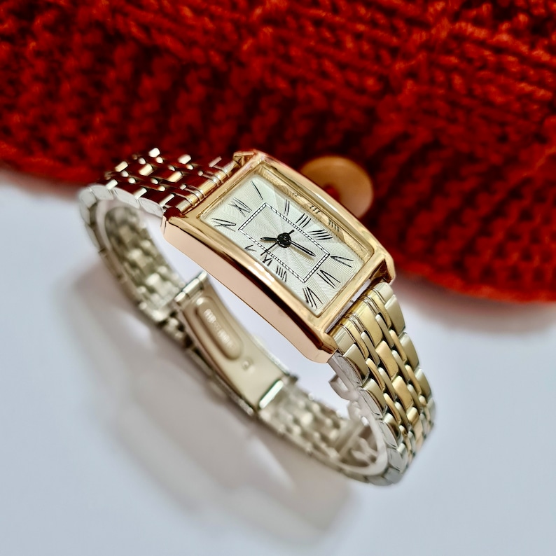 May include: A gold and silver toned watch with a rectangular face and Roman numeral hour markers. The watch has a two-tone metal band.