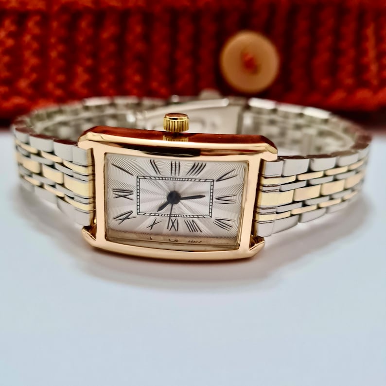 May include: A gold and silver two-tone watch with a square face and Roman numeral hour markers. The watch has a silver band with gold accents.