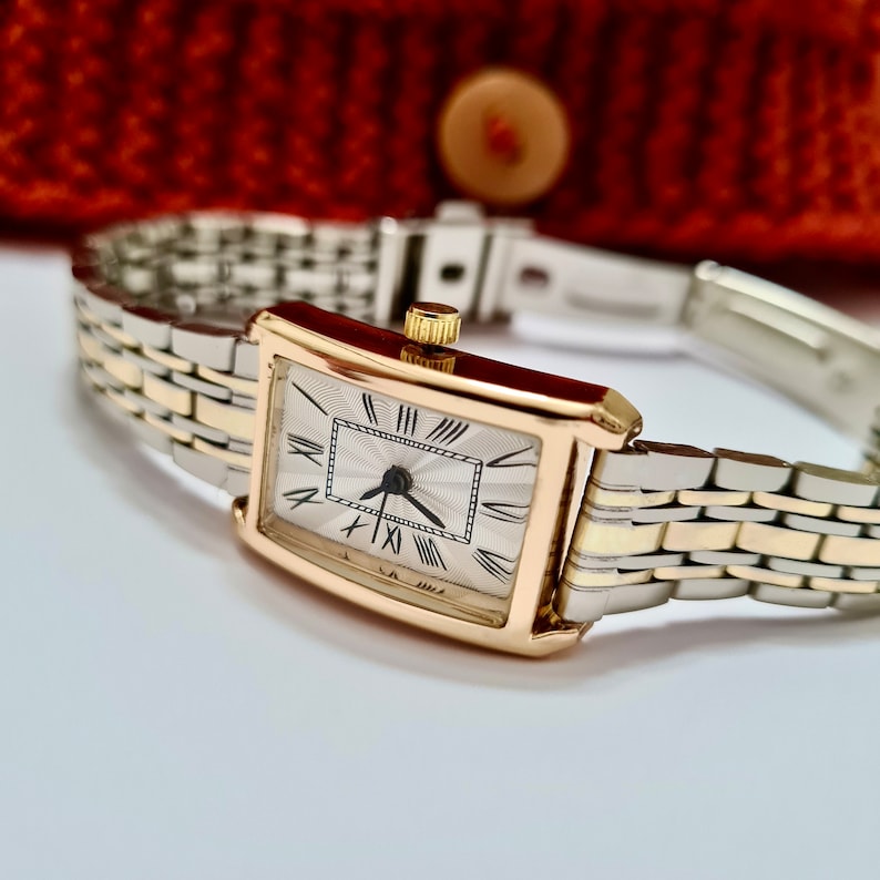 May include: A gold and silver toned wristwatch with a rectangular face and Roman numeral hour markers. The watch has a white face with a silver-toned hour and minute hand and a black second hand.