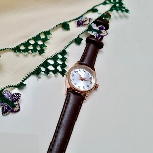 May include: A brown leather wristwatch with a rose gold case and white face. The watch has black hour and minute hands and black Roman numeral hour markers. The watch is lying on a white surface with a green and white lace fabric.