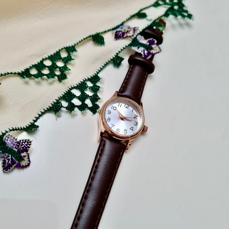 May include: A brown leather wristwatch with a rose gold case and white face. The watch has black hour and minute hands and black Roman numeral hour markers. The watch is lying on a white surface with a green and white lace fabric.