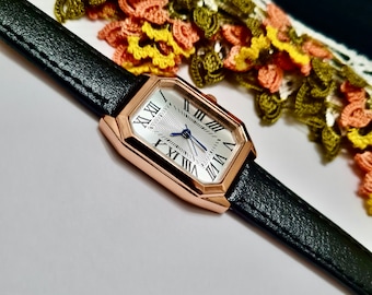 Op de afbeelding: Een roségouden horloge met een zwarte leren band en een witte wijzerplaat met Romeinse cijfers. Het horloge heeft een vierkante wijzerplaat met afgeronde hoeken.
