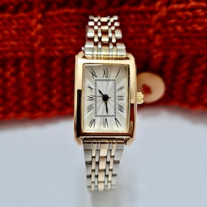 May include: A gold-tone watch with a rectangular face and Roman numeral hour markers. The watch has a two-tone silver and gold metal band.