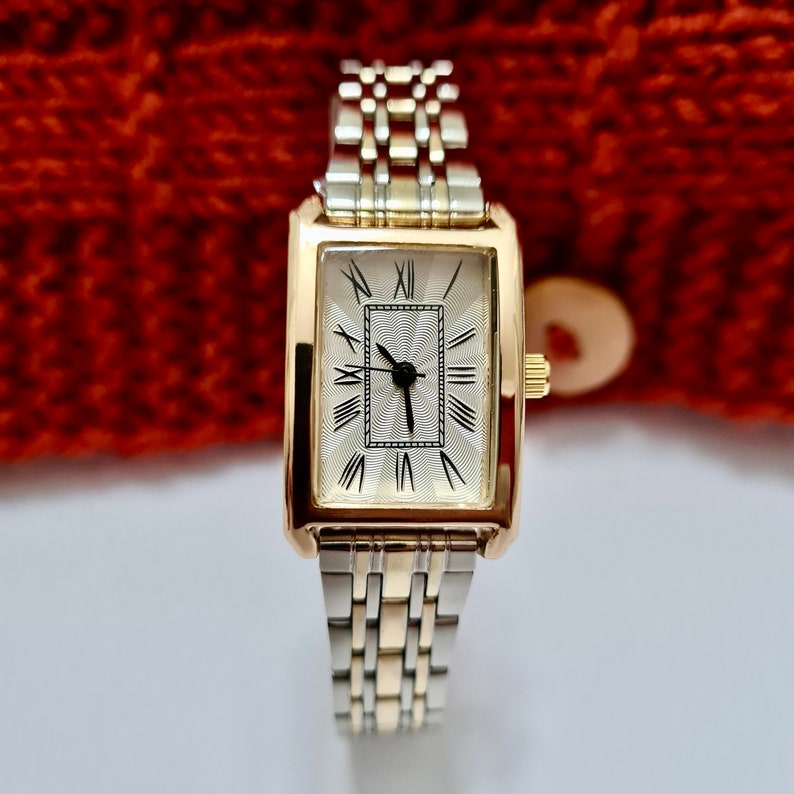 May include: A gold-tone watch with a rectangular face and Roman numeral hour markers. The watch has a two-tone silver and gold metal band.