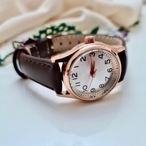 May include: A rose gold wristwatch with a white face and black numbers. The watch has a brown leather band.