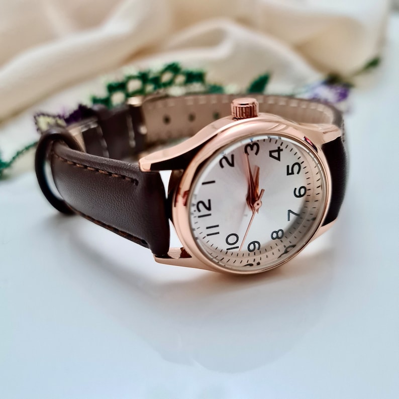 May include: A rose gold wristwatch with a white face and black numbers. The watch has a brown leather band.