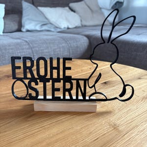 Puede incluir: Decoración de Pascua de metal negro con el texto alemán "FROHE OSTERN" y una silueta de conejo. Montada sobre una pequeña base rectangular de madera, perfecta para la mesa.