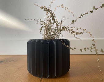 Puede incluir: Un macetero cilíndrico negro con diseño acanalado, que contiene una planta de helecho seca.