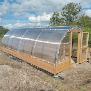 Peut inclure: Une serre à ossature en bois avec un toit en plastique transparent et une porte en bois. La serre est posée sur un sol en terre.