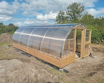 May include: A wooden framed greenhouse with a clear plastic roof and a wooden door. The greenhouse is sitting on a dirt ground.
