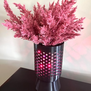 May include: A black vase with a geometric pattern and a pink flower arrangement. The vase has a light inside that illuminates the flowers.