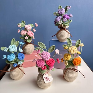 Op de afbeelding: Een verzameling handgemaakte gehaakte bloemboeketten in kleine beige vazen. De bloemen zijn in verschillende kleuren, waaronder roze, blauw, geel en paars. Elke vaas is vastgebonden met een bruin lint. Eén vaas heeft een hartvormig label met "Happy Valentine's Day".