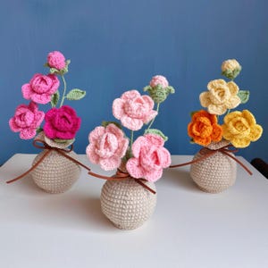 Puede incluir: Tres pequeños jarrones de ganchillo sostienen cada uno un ramo de flores. Las flores son en tonos de rosa, naranja y amarillo, con hojas verdes. Los jarrones son beige y están atados con cintas marrones. El fondo es azul liso.