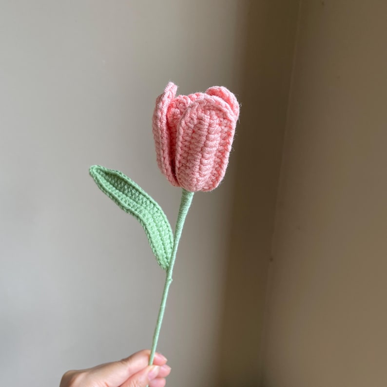 May include: A handmade crochet tulip in shades of pink, green, and light green. The flower has a detailed, textured appearance, with a green stem and leaf. The tulip is held by a person.