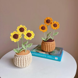 Op de afbeelding: Twee gehaakte zonnebloemarrangementen in kleine potten. Eén pot is beige, de andere is oranje. Elke zonnebloem heeft gele bloemblaadjes en een bruin hart. De potten staan op een witte tafel, met een boek ertussen.