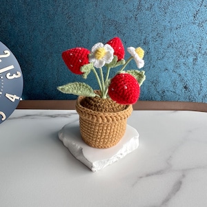Op de afbeelding: Een gehaakte aardbeienplant in een bruine pot. De plant heeft felrode aardbeien, witte bloemen met gele centra en groene bladeren. De pot staat op een witte, onregelmatig gevormde basis.