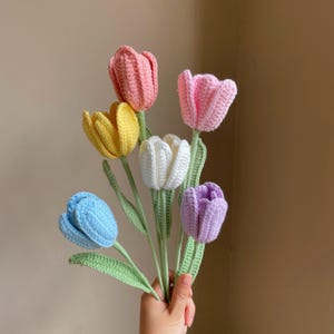 May include: A bouquet of handmade crochet tulips in various colors, including pink, yellow, white, blue, and purple. The flowers have green stems and leaves. The tulips are held in a hand.