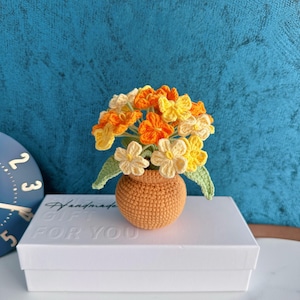 Peut inclure: Un bouquet crocheté de fleurs jaunes, oranges et crème dans un petit vase crocheté orange. Le vase est posé sur une boîte blanche avec les mots "Handmade Gift For You". Le fond est de couleur bleu-vert texturé.