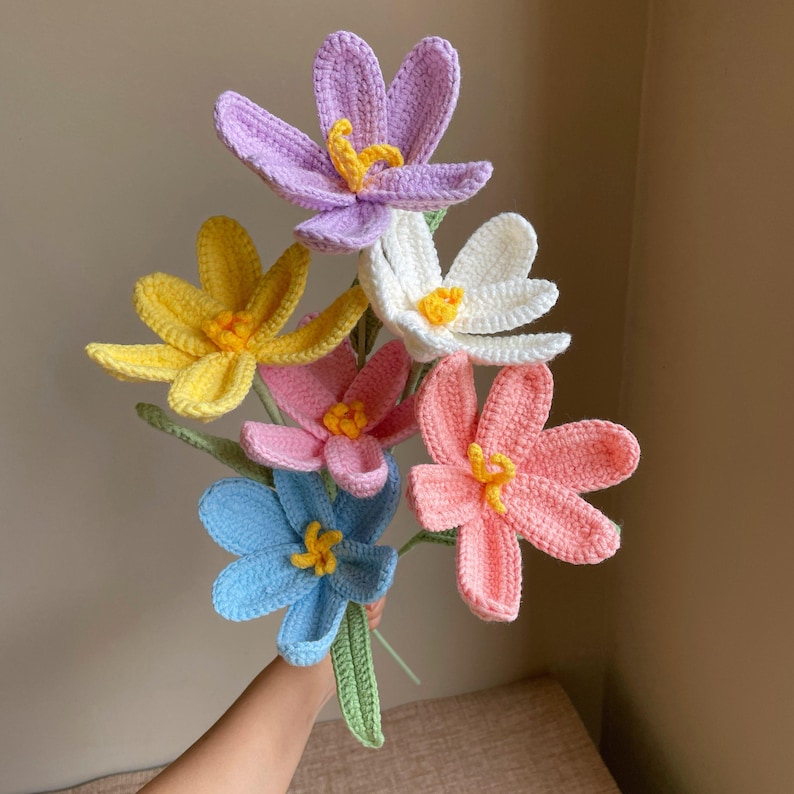 May include: A bouquet of handmade crochet flowers in various colors, including purple, yellow, white, pink, blue, and peach. The flowers have yellow centers and green stems, creating a vibrant and cheerful arrangement.