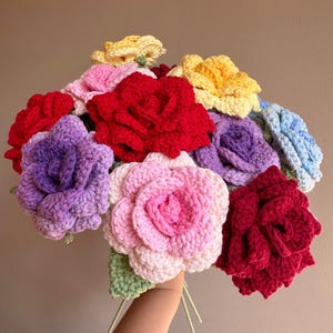 May include: A colourful bouquet of crocheted roses in shades of red, pink, yellow, purple, and blue. The handmade flowers are held together with green stems and leaves, creating a textured and vibrant floral arrangement.