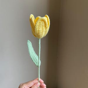 May include: A handmade crochet tulip in bright yellow with a green stem and leaf. The flower's petals are tightly closed, and the leaf is a soft green. The tulip is a charming, handcrafted item.