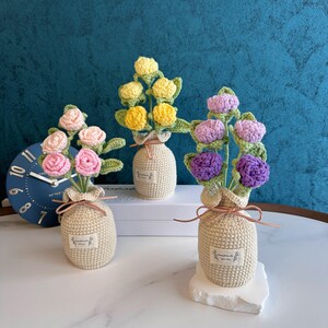 Pode incluir: Três arranjos de flores de crochê em vasos bege. As flores são rosa, amarelas e roxas, com folhas verdes. Cada vaso tem uma pequena etiqueta e é amarrado com uma fita marrom. Os arranjos estão em uma superfície branca.