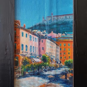 May include: Oil painting of a European street scene in a black frame. The artwork depicts colorful buildings with a blue sky backdrop. The buildings are painted in shades of orange, pink, and white, with green trees lining the street.