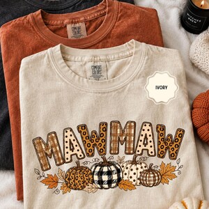 May include: An ivory t-shirt featuring the text MAWMAW in a mix of plaid, leopard, and polka dot patterns, with a graphic of four pumpkins and autumn leaves below. The shirt has a Comfort Colors label at the neck and is layered with other shirts.