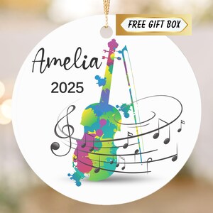 May include: A white round ornament with a colorful violin design, musical notes, and the name "Amelia" and the year "2025". The violin is decorated with splashes of blue, green, pink, and yellow. A gold ribbon is attached for hanging.