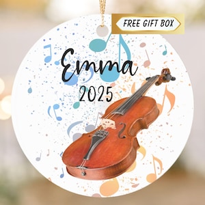 May include: A round, white ornament featuring a watercolor-style violin with musical notes in blue and orange. The name "Emma" and the year "2025" are written on the ornament. A gold ribbon is attached for hanging.