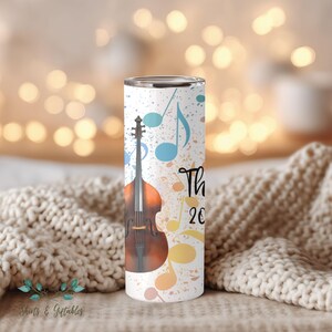 May include: A white tumbler featuring a brown upright bass and musical notes in blue, yellow, and orange. The text "Thank 20" is written in black script. The tumbler has a stainless steel rim and is decorated with a speckled background.