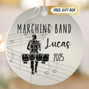 May include: A round, white ornament with a gold string for hanging. The ornament features a black illustration of a marching band drummer, musical notes, and the text "MARCHING BAND Lucas 2025". A gold banner reads "FREE GIFT BOX".