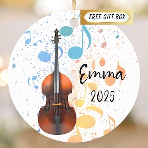 May include: A white, round ornament featuring a brown double bass with musical notes in blue, yellow, and orange. The name "Emma" and the year "2025" are written in black script. The words "FREE GIFT BOX" are in a gold banner.