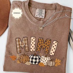 May include: An espresso-colored short-sleeve t-shirt with a crew neckline. The graphic features the word MIMI in mixed patterns above four pumpkins with leopard, buffalo plaid, and polka dot prints, surrounded by orange and brown autumn leaves.
