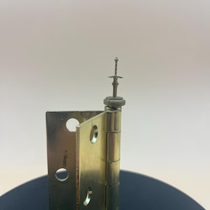 May include: A brass-colored hinge with a decorative sword-shaped element on top. The hinge is open, revealing the holes for screws. The background is a neutral color.