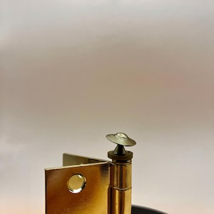 May include: A close-up of a gold-coloured hinge with a decorative top. The hinge is made of metal and has a shiny finish. The hinge is attached to a black surface. The background is a gradient of white and beige.