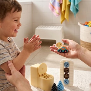 May include: A beige plastic potty-shaped toy, a white rectangular frame with four circular icons, a small orange and blue toy car, a square card with a yellow smiley face, and two small cone-shaped toys in black and blue.